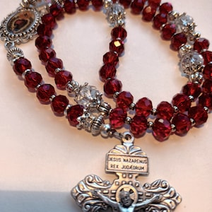 May include: A rosary with deep red faceted beads and silver-toned metal accents. The rosary features a crucifix with the inscription "JESUS NAZARENUS REX JUDAEORUM" and small religious icons.