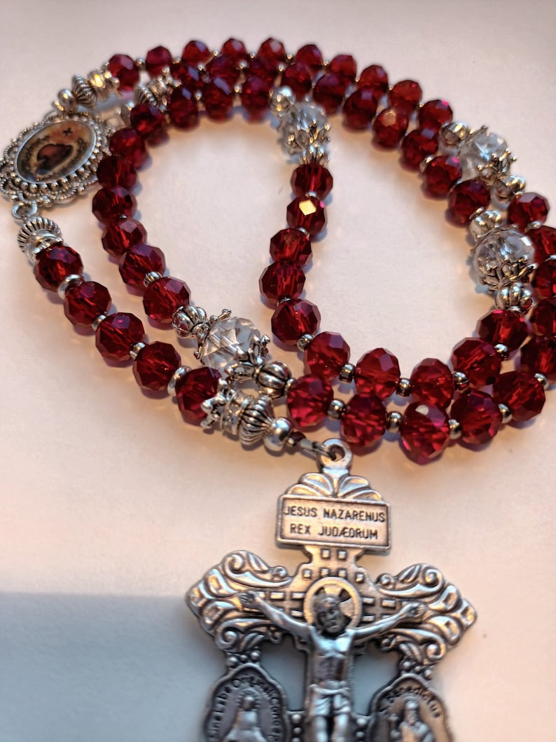 May include: A rosary with deep red faceted beads and silver-toned metal accents. The rosary features a crucifix with the inscription "JESUS NAZARENUS REX JUDAEORUM" and small religious icons.