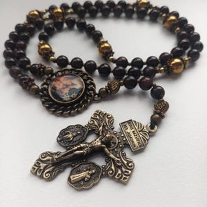 May include: A rosary with dark brown beads and ornate bronze-colored metal accents. The centerpiece features a small image, and the crucifix displays a figure of Jesus with the inscription "Jesus Nazarenus Rex Judaeorum."