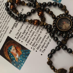 May include: A rosary featuring dark gray beads, metallic bronze-colored accent beads, and ornate bronze-toned metal spacers. The rosary includes a circular pendant containing a small image of a heart with rays of light emanating from it.