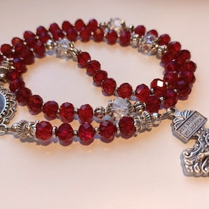 May include: A rosary with deep red faceted beads and silver-toned metal accents. The rosary features a decorative pendant with a small image and a detailed crucifix with the inscription "Jesus Nazarenus Rex Judaeorum".