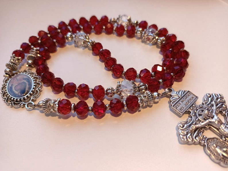May include: A rosary with deep red faceted beads and silver-toned metal accents. The rosary features a decorative pendant with a small image and a detailed crucifix with the inscription "Jesus Nazarenus Rex Judaeorum".