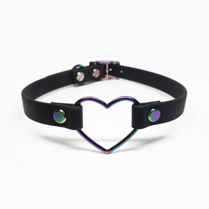 Może przedstawiać: Czarny choker z metalowym pierścieniem w kształcie tęczowego serca. Choker ma po dwie czarne zatrzaski po obu stronach.