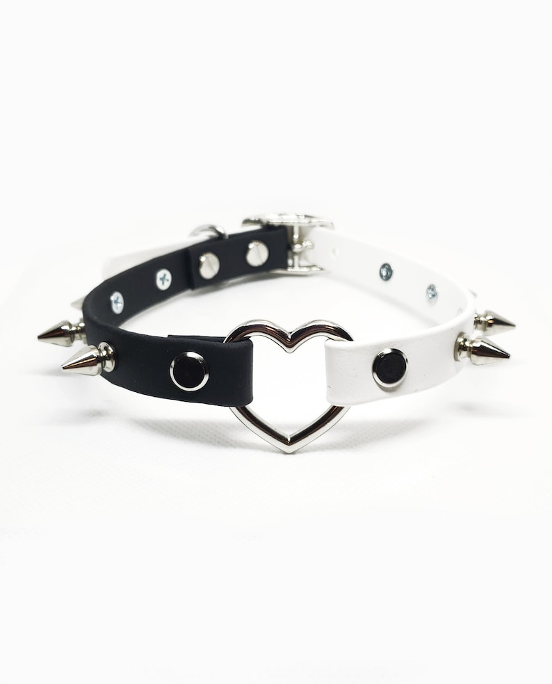 Op de afbeelding: Een zwart-witte choker van imitatieleer met zilveren spikes en een zilveren ring in de vorm van een hart.