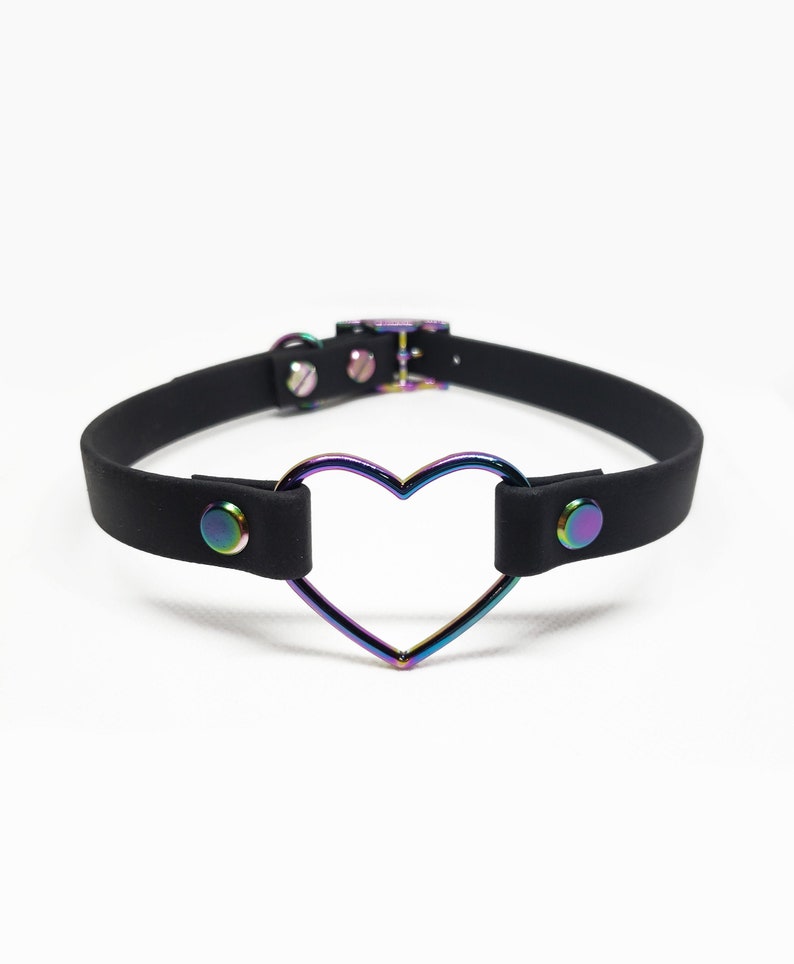 Może przedstawiać: Czarny choker z metalowym pierścieniem w kształcie tęczowego serca. Choker ma po dwie czarne zatrzaski po obu stronach.