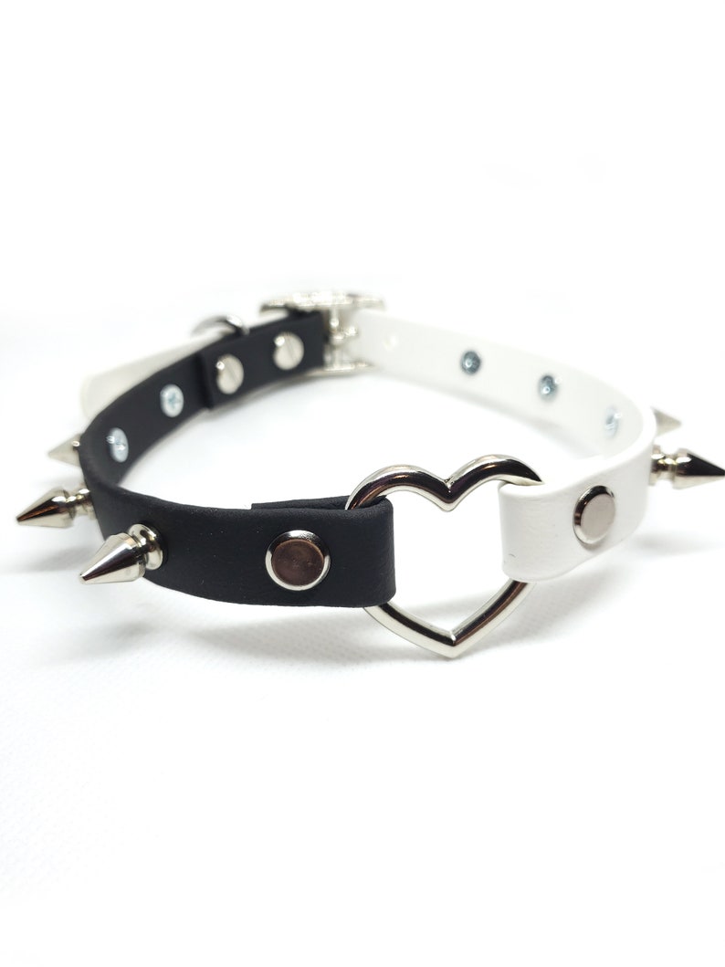 Op de afbeelding: Een zwart-witte choker met een hartvormige metalen ring en zilveren spikes. De choker is gemaakt van imitatieleer.