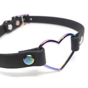 Może przedstawiać: Czarny sk&oacute;rzany choker z metalowym pierścieniem w kształcie tęczowego serca. Choker ma zapięcie na klamrę i dwa srebrne nity.