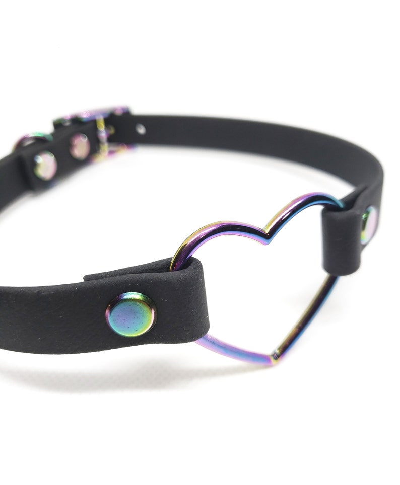 Może przedstawiać: Czarny sk&oacute;rzany choker z metalowym pierścieniem w kształcie tęczowego serca. Choker ma zapięcie na klamrę i dwa srebrne nity.