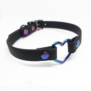 Może przedstawiać: Czarny choker z metalowym pierścieniem w kształcie tęczowego serca. Choker ma zapięcie na klamrę i jest ozdobiony srebrnymi ćwiekami.
