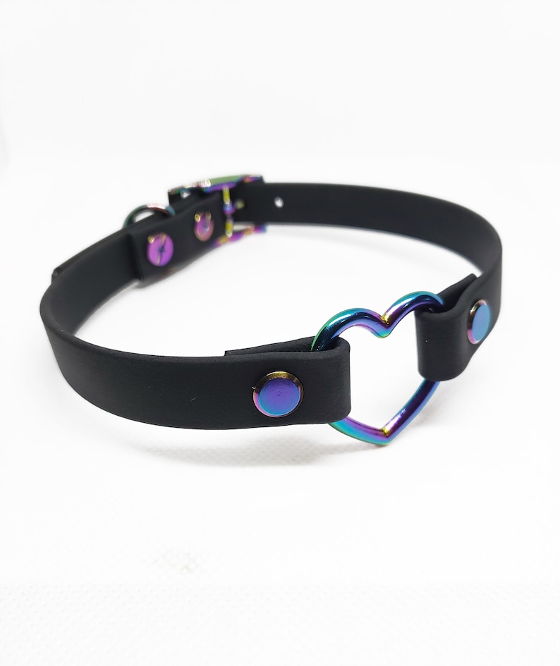 Może przedstawiać: Czarny choker z metalowym pierścieniem w kształcie tęczowego serca. Choker ma zapięcie na klamrę i jest ozdobiony srebrnymi ćwiekami.