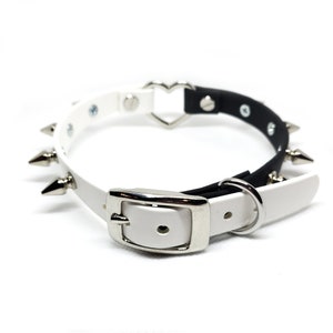 Op de afbeelding: Een witte en zwarte leren choker met zilveren spikes en een hartvormige gesp. De choker is verstelbaar met een zilveren gesp en een zilveren ring.