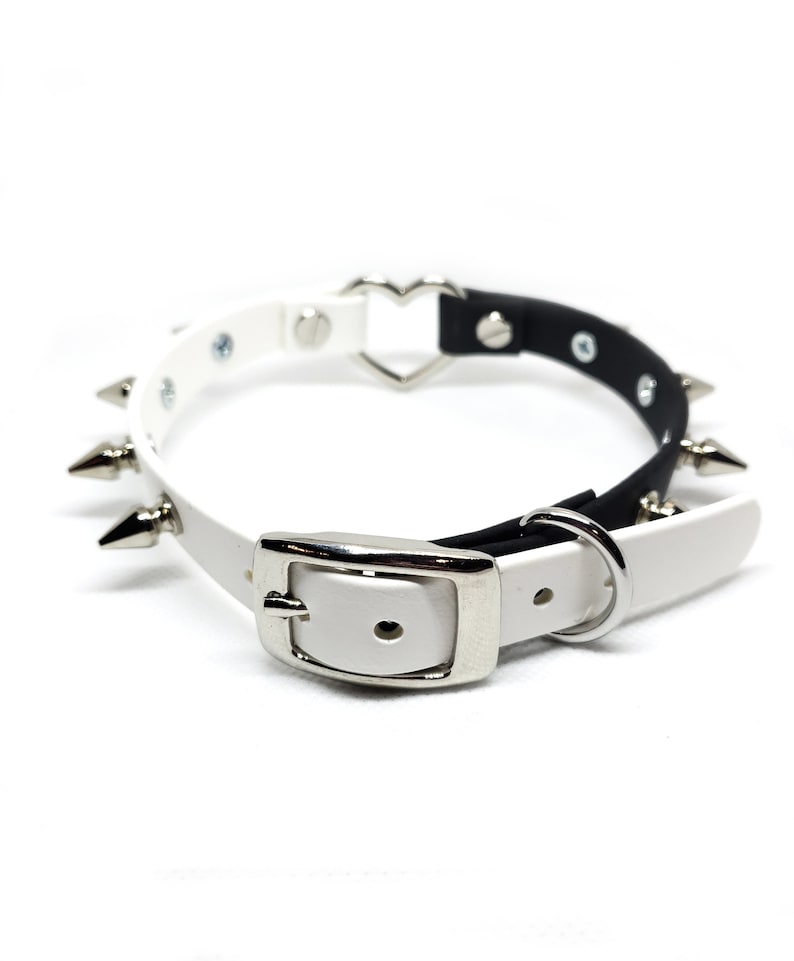 Op de afbeelding: Een witte en zwarte leren choker met zilveren spikes en een hartvormige gesp. De choker is verstelbaar met een zilveren gesp en een zilveren ring.