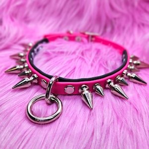May include: A hot pink collar with silver-colored cone studs and a large metal ring. The collar is set against a fluffy pink background. The collar is a fashion accessory.