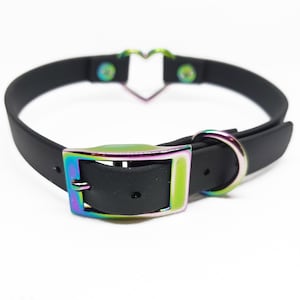 Może przedstawiać: Czarny choker z metalowym akcentem w kształcie serca i tęczową, opalizującą klamrą.