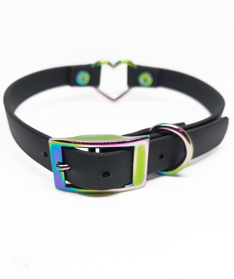 Może przedstawiać: Czarny choker z metalowym akcentem w kształcie serca i tęczową, opalizującą klamrą.