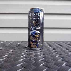 Puede incluir: Una lata negra de Guinness Draught Stout con el texto "Gives America Back" y un gr&aacute;fico de un p&aacute;jaro azul volando sobre un paisaje con una autocaravana.