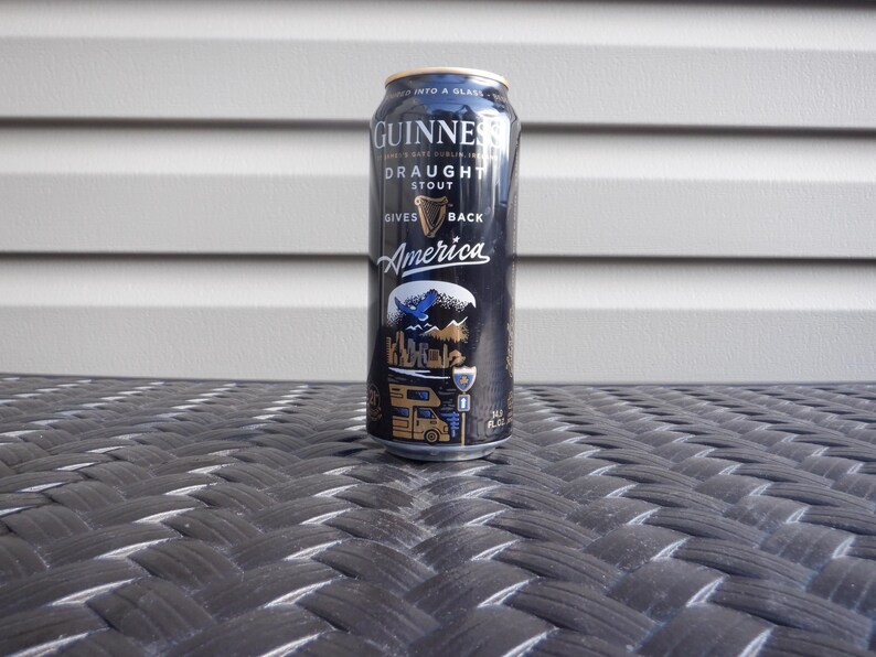 Puede incluir: Una lata negra de Guinness Draught Stout con el texto "Gives America Back" y un gr&aacute;fico de un p&aacute;jaro azul volando sobre un paisaje con una autocaravana.