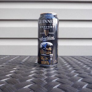 Puede incluir: Una lata negra de Guinness Draught Stout con el texto "Gives America Back" y un gr&aacute;fico de una autocaravana conduciendo por un paisaje pintoresco.