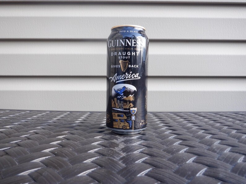Puede incluir: Una lata negra de Guinness Draught Stout con el texto "Gives America Back" y un gr&aacute;fico de una autocaravana conduciendo por un paisaje pintoresco.