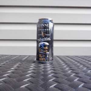 Puede incluir: Una lata negra de Guinness Draught Stout con una etiqueta dorada. La etiqueta dice "Gives America Back" y presenta un gr&aacute;fico de un p&aacute;jaro azul y una autocaravana.