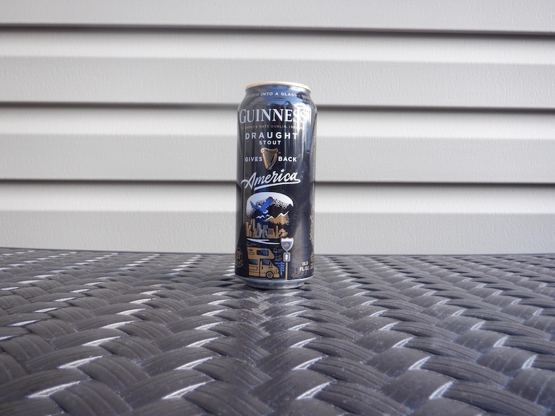 Puede incluir: Una lata negra de Guinness Draught Stout con una etiqueta dorada. La etiqueta dice "Gives America Back" y presenta un gr&aacute;fico de un p&aacute;jaro azul y una autocaravana.
