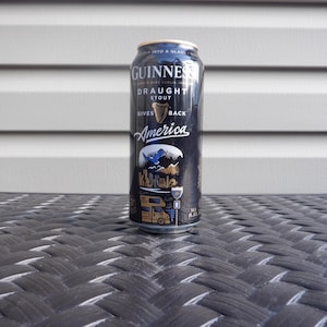 Puede incluir: Una lata negra de Guinness Draught Stout con letras doradas. La lata tiene un dise&ntilde;o con un p&aacute;jaro azul, monta&ntilde;as y una autocaravana. El texto en la lata dice "Gives Back America".