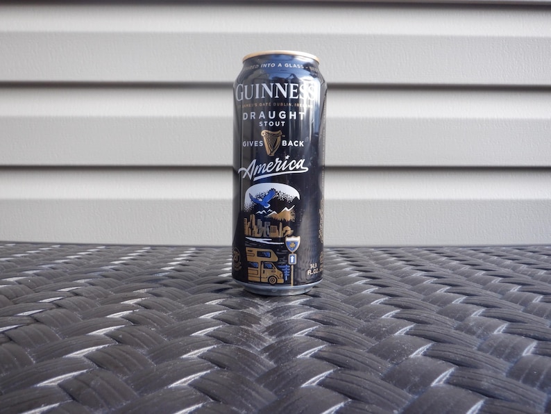 Puede incluir: Una lata negra de Guinness Draught Stout con letras doradas. La lata tiene un dise&ntilde;o con un p&aacute;jaro azul, monta&ntilde;as y una autocaravana. El texto en la lata dice "Gives Back America".