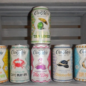 May include: Six different aluminum cans of Cape May Brewing Co. beer. The cans are arranged in two rows of three. The top row features a can of Tan Limes, a Mexican-style lager with sea salt and lime. The bottom row features cans of Citrus Shandy The Grove, India Pale Ale Cape May IPA, Cranberry Shandy The Bog, Marzen-Style Lager Oktoberfest, and Cape May White.