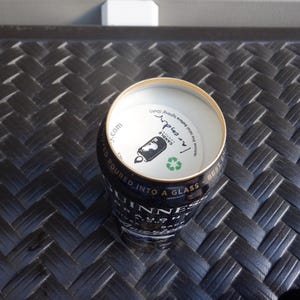 Puede incluir: Una lata plateada con una etiqueta negra y dorada que dice "Guinness" y "Draught" con un s&iacute;mbolo de reciclaje verde. La parte superior de la lata est&aacute; abierta y muestra una etiqueta blanca con el texto "I'm inside" y "Canola", con un gr&aacute;fico en blanco y negro de una lata.