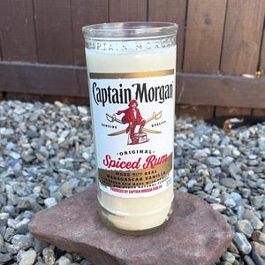 May include: A clear glass bottle of Captain Morgan Spiced Rum, filled with a light-colored liquid. The label features a red-coated pirate and the words "Spiced Rum." The bottle is resting on a rock.