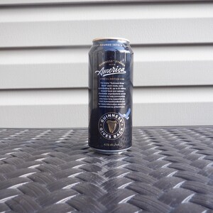 Puede incluir: Una lata negra de cerveza Guinness con el texto "Guinness Stands with America" y "Special Edition Can". La lata tambi&eacute;n tiene el texto "Guinness Gives Back" y un gr&aacute;fico de un p&aacute;jaro azul.