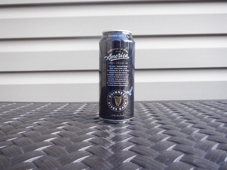 Puede incluir: Una lata negra de cerveza Guinness con el texto "Guinness Stands with America" y "Special Edition Can". La lata tambi&eacute;n tiene el texto "Guinness Gives Back" y un gr&aacute;fico de un p&aacute;jaro azul.