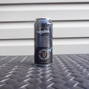Puede incluir: Una lata negra de cerveza Guinness con las palabras "Guinness Stands with America" y "Guinness Gives Back" impresas en ella. La lata est&aacute; sobre una superficie de mimbre negra.