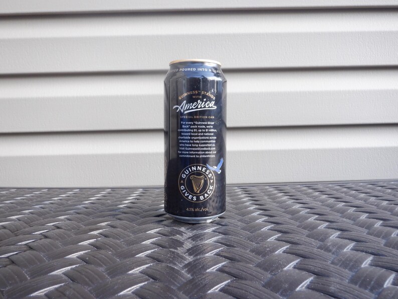 Puede incluir: Una lata negra de cerveza Guinness con las palabras "Guinness Stands with America" y "Guinness Gives Back" impresas en ella. La lata est&aacute; sobre una superficie de mimbre negra.