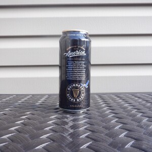 Puede incluir: Una lata negra de cerveza Guinness con el texto "Guinness Stands with America" y "Guinness Gives Back".