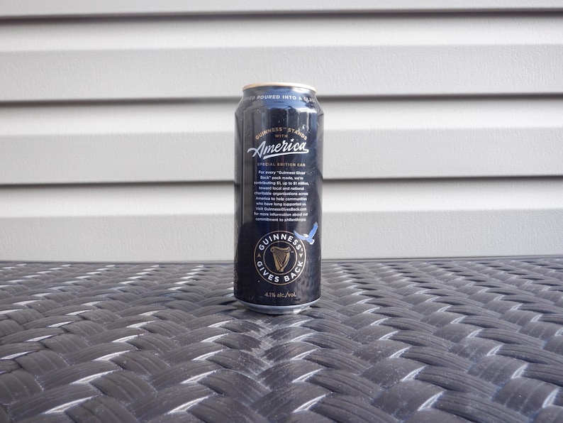 Puede incluir: Una lata negra de cerveza Guinness con el texto "Guinness Stands with America" y "Guinness Gives Back".