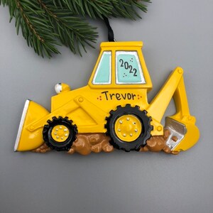 May include: Yellow toy backhoe Christmas ornament with the name "Trevor" and the year "2022" written on it.