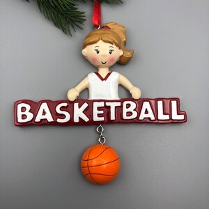 May include: A basketball-themed ornament featuring a female figure in a white and maroon basketball uniform. The ornament has the word "BASKETBALL" in white letters on a maroon banner. A small orange basketball hangs below.