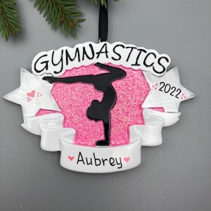 May include: Pink and white Christmas ornament with a black silhouette of a gymnast performing a handstand. The ornament has the word "Gymnastics" and the year "2022" on it. The name "Aubrey" is written on a white banner.