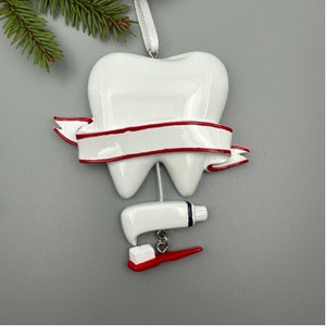 May include: White tooth-shaped Christmas ornament with a red ribbon and a blank banner. The ornament also features a white tube of toothpaste and a red toothbrush.