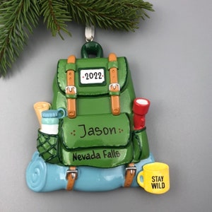 Könnte beinhalten: Grüner Rucksack-Weihnachtsbaumschmuck mit braunem Gurt, einem blauen Schlafsack, einer roten Taschenlampe, einer Wasserflasche, einer Karte und einer gelben Tasse mit der Aufschrift "Stay Wild". Der Schmuck ist mit dem Namen "Jason" und dem Text "Nevada Falls" sowie dem Jahr "2022" personalisiert.