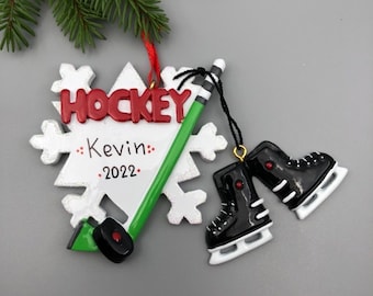 May include: A white snowflake ornament with the word "HOCKEY" in red letters, a green and black hockey stick, and the text "Kevin 2022". Two black ice skates are attached to the ornament with black string.