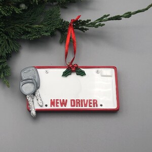 May include: A white ceramic ornament shaped like a license plate with red trim. The ornament has a holly sprig with red berries, two silver keys, and the text "NEW DRIVER." hanging from the top is a red ribbon.