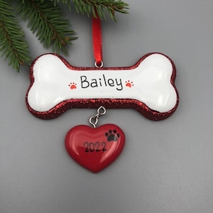 May include: White bone-shaped Christmas ornament with red glitter trim and the name "Bailey" written on it. A red heart with a paw print and the year "2022" is hanging from the bone.