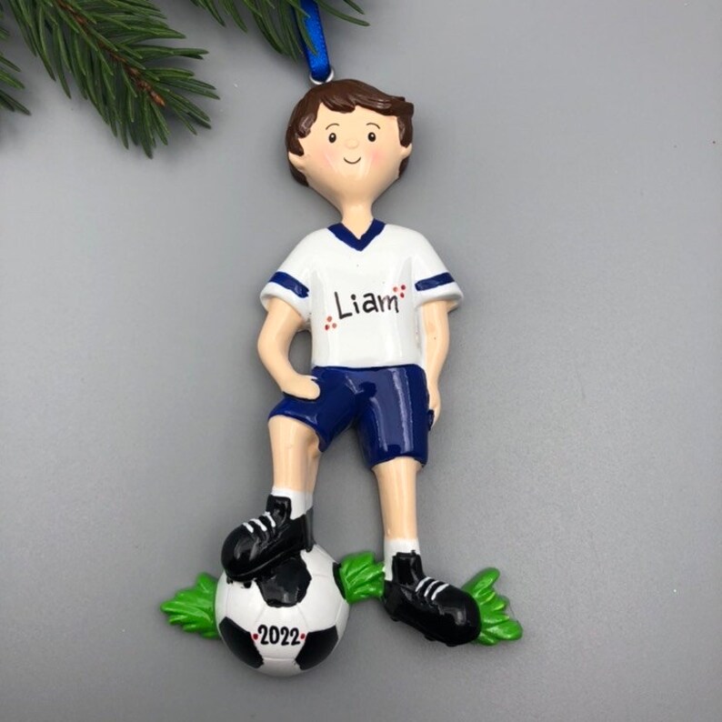 May include: Personalized Christmas ornament featuring a cartoon boy soccer player with brown hair, wearing a white jersey with blue trim, blue shorts, and black cleats. The ornament includes the name "Liam" on the jersey and the year "2022" on the soccer ball.