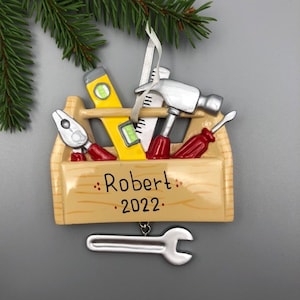 May include: A wooden toolbox Christmas ornament with a hammer, screwdriver, pliers, and level. The toolbox is decorated with the name "Robert" and the year "2022". A silver wrench hangs from the ornament.
