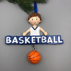 Pu&ograve; includere: Un ornamento da basket bianco e blu con un ragazzo che indossa una maglia bianca e pantaloncini blu. L'ornamento ha la scritta "BASKETBALL" in lettere bianche. Un piccolo pallone da basket arancione &egrave; appeso all'ornamento.