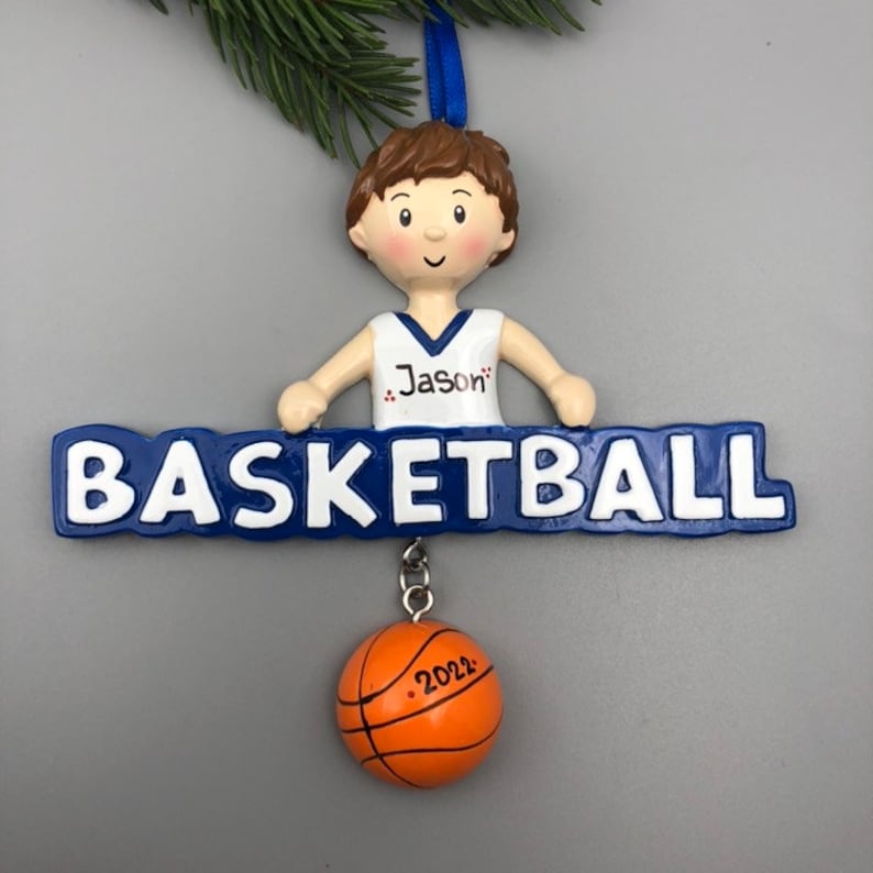 Pu&ograve; includere: Un ornamento natalizio personalizzato blu e bianco con un ragazzo in uniforme da basket che tiene uno striscione con la scritta "BASKETBALL" e il nome "Jason" sull'uniforme. Un pallone da basket con l'anno "2022" &egrave; appeso allo striscione.