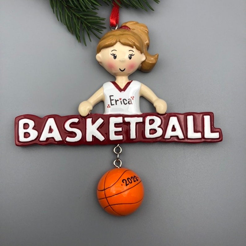 May include: A basketball-themed ornament featuring a female figure in a white and maroon basketball jersey with the name "Erica". The ornament has a maroon banner that reads "BASKETBALL" and a small orange basketball with "2022" on it.