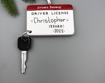 May include: White and red Christmas ornament shaped like a driver's license with the text "Drivers Beware!" and "DRIVER LICENSE" and the name "Christopher" written in black marker. The ornament also includes the text "ISSUED: 2022". A silver key with a black head is attached to the ornament.